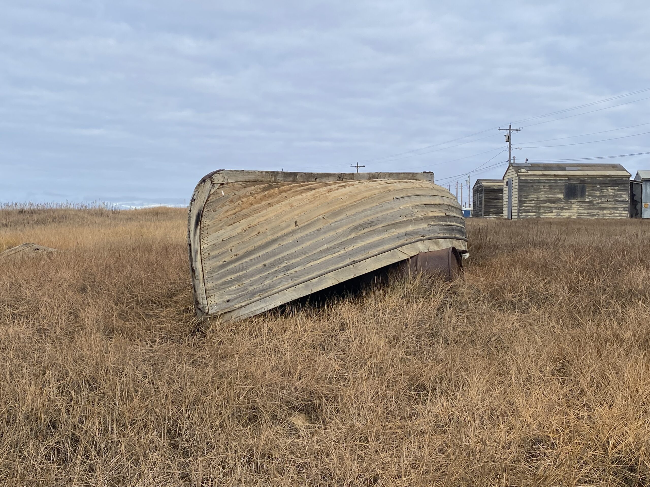 Image of a weathered wooden boat upside-down in the grass with wooden buildings in the background.