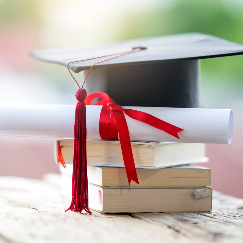 Graduation cap sitting atop a stack of books and a rolled up degree tied with a red ribbon.