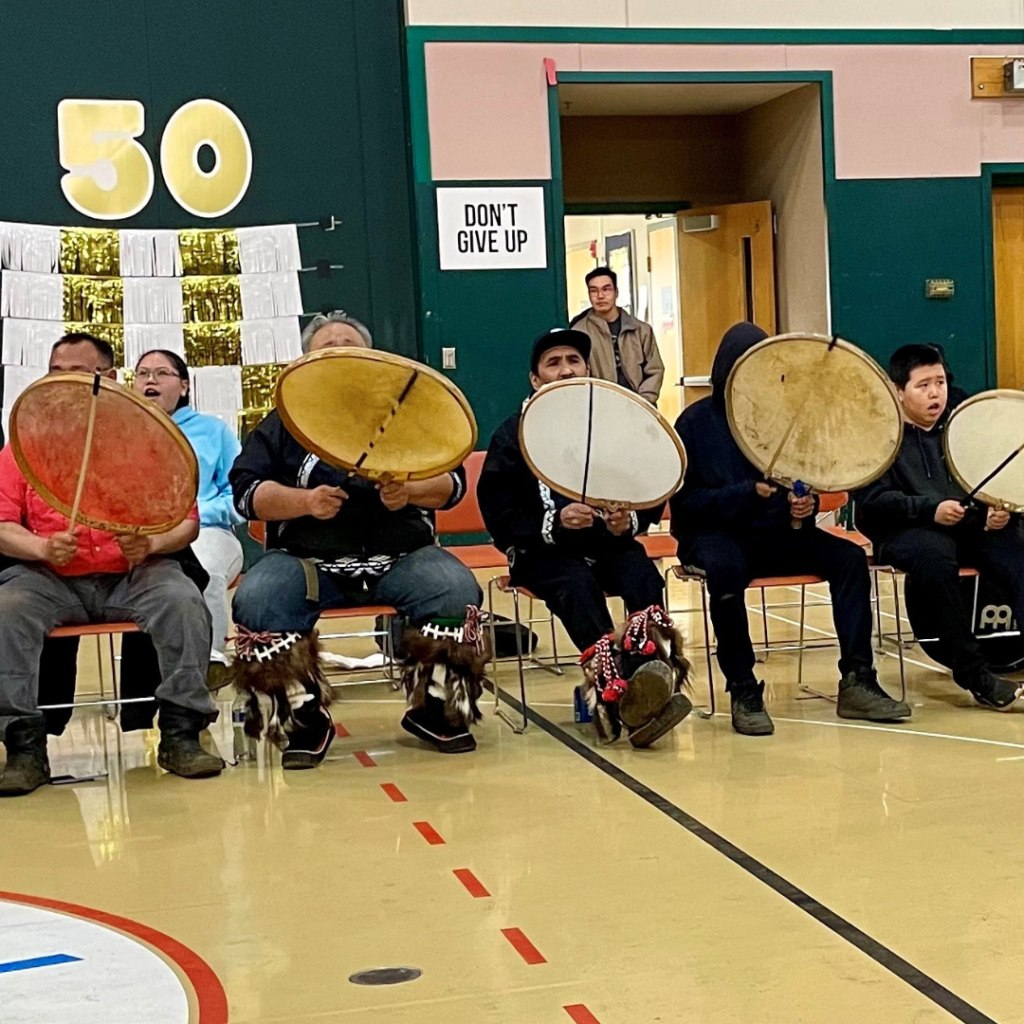 Group of Alaska Native drummers playing traditional drums in a school gym.